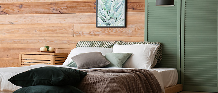 Schlafzimmereinrichtung mit Holzwand, Massivholzbett und hölzernem Nachttisch, dazu Kissen und Decken in natürlichen Farben wie Grün und Braun