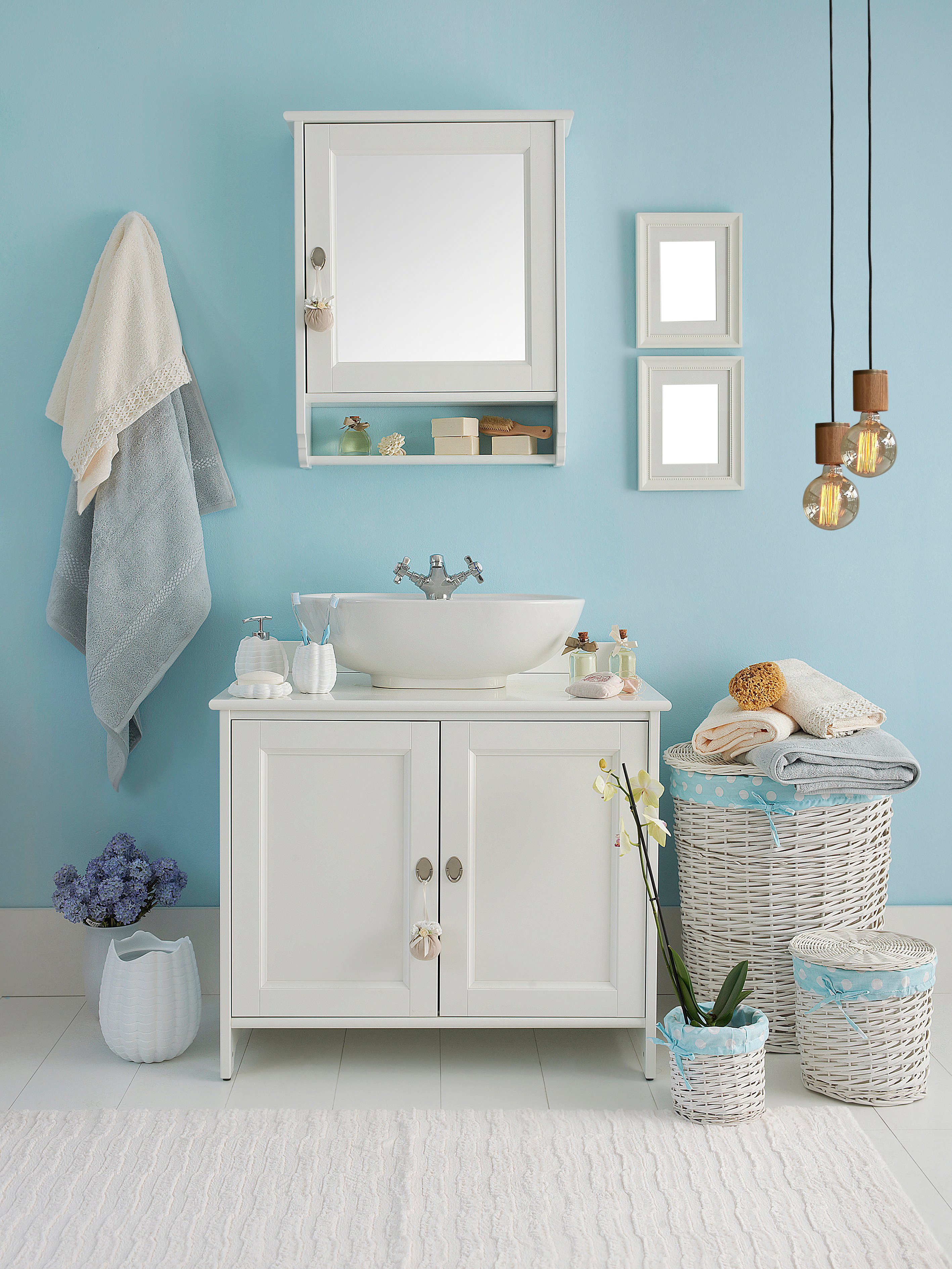 Eine Unterschrank eines Waschbeckens und der passende Wandspiegel sind an einer blauen Badezimmerwand angebracht. Links und Rechts sind mehrere Deko- Elemente.