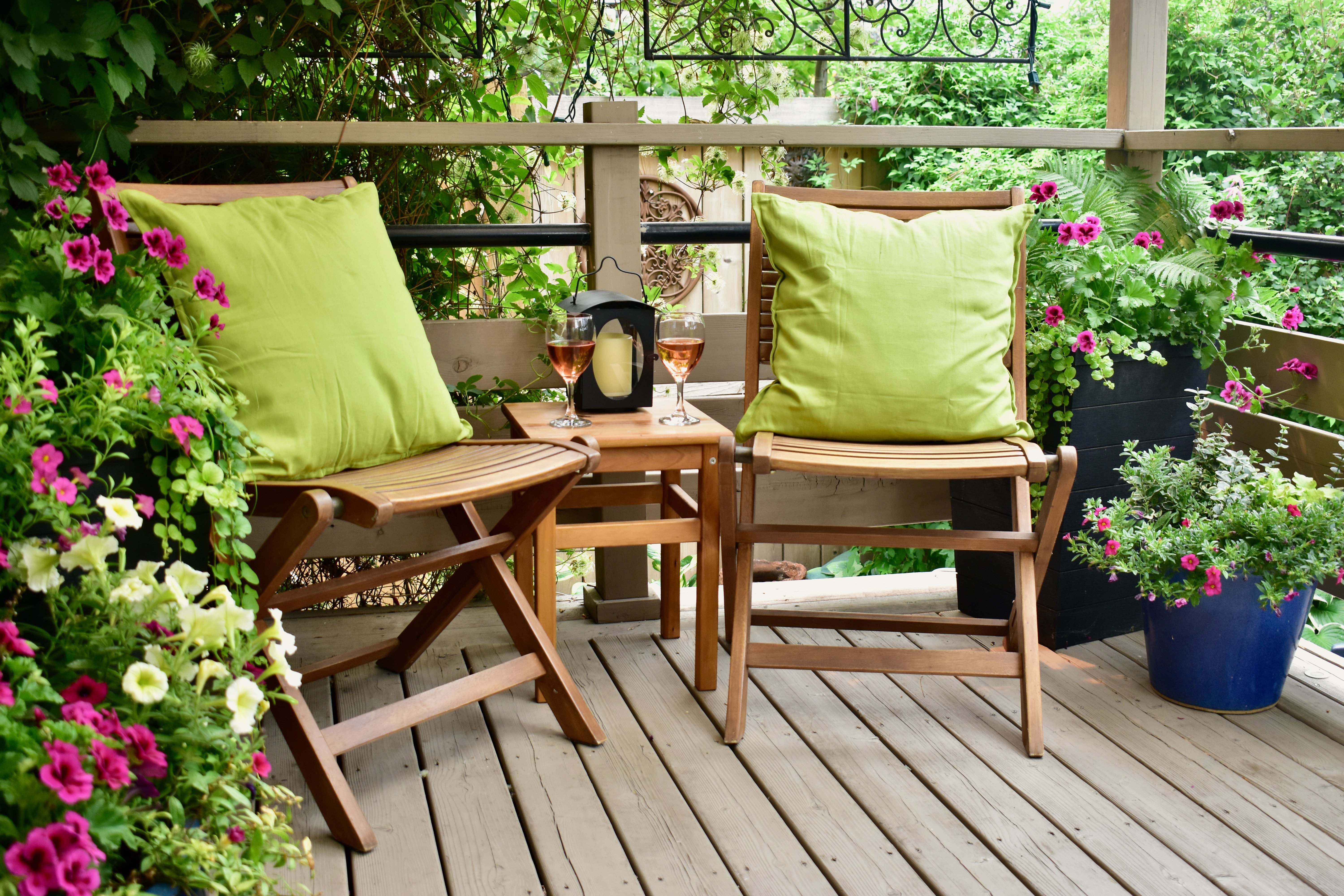 Zwei Gartenstühle aus Holz stehen auf einen Balkon. Auf den Stühlen befindet sich jeweils ein grünes Kissen. Links und rechts neben den Stühlen stehen Blumen.