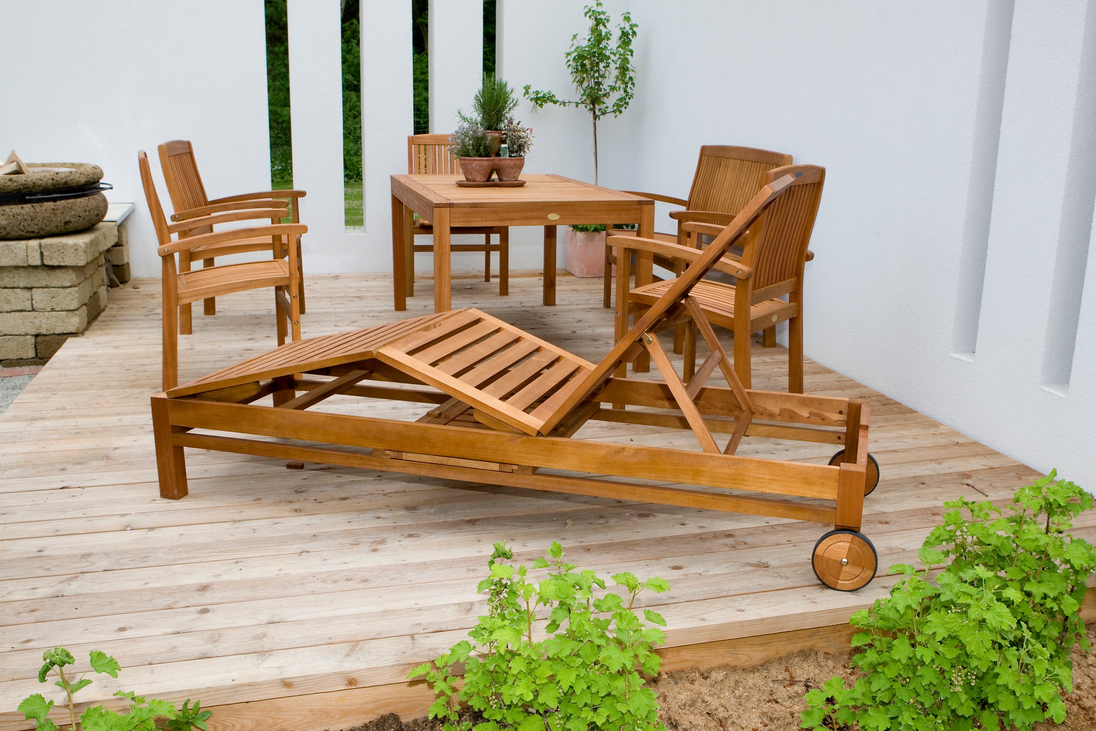 Eine Gartenliege aus Holz steht auf einer Terrasse. Im Hintergrund befinden sich passende Gartenstühle und ein Gartentsich.
