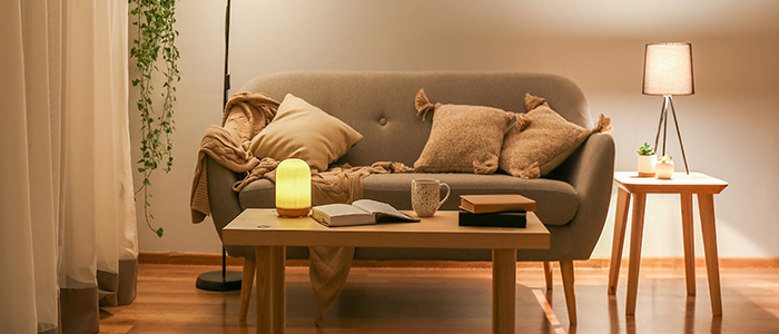 Wohnzimmereinrichtung in Abendstimmung mit grauem Sofa, hellen Gardinen, Holztischen, beigem Teppich sowie Deckenfluter und Tischlampe